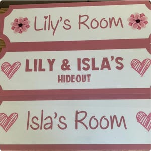 Puede incluir: Tres letreros rectangulares en rosa y blanco. El letrero superior dice "Lily's Room" con una flor rosa. El letrero del medio dice "LILY & ISLA'S HIDE OUT" con un coraz&oacute;n. El letrero inferior dice "Isla's Room" con un coraz&oacute;n.