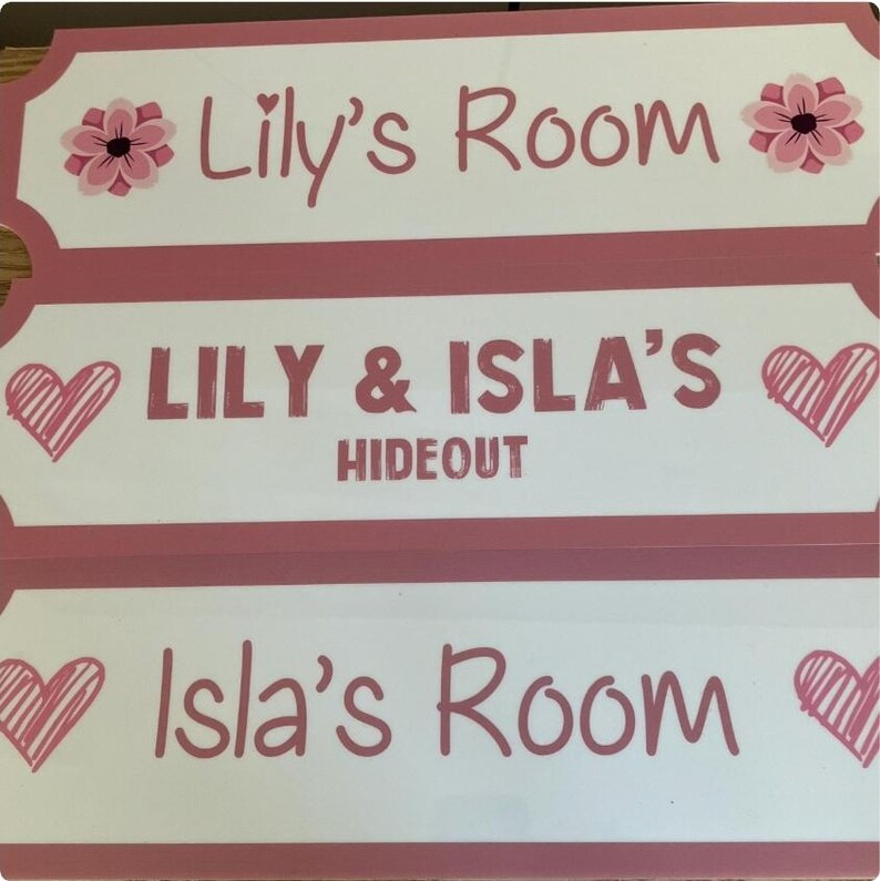 Puede incluir: Tres letreros rectangulares en rosa y blanco. El letrero superior dice "Lily's Room" con una flor rosa. El letrero del medio dice "LILY & ISLA'S HIDE OUT" con un coraz&oacute;n. El letrero inferior dice "Isla's Room" con un coraz&oacute;n.
