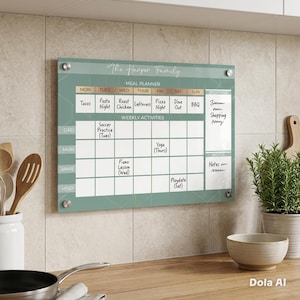 May include: A rectangular wall-mounted acrylic meal planner and weekly activity calendar in sage green with gold geometric lines. It features the text The Harper Family, columns for days of the week, rows for family members, and sections for shopping and notes.