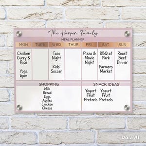 May include: A rectangular acrylic meal planner board with a pale pink background and rose gold accents. It features the text The Harper Family Meal Planner, days of the week, and sections for shopping and snack ideas with handwritten black text.