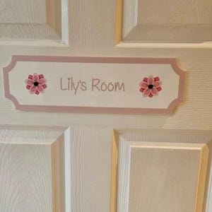 Puede incluir: Un letrero rectangular rosa claro con el texto "Lily's Room" en cursiva. El letrero est&aacute; decorado con dise&ntilde;os florales rosas y rojos. Est&aacute; montado en una puerta blanca.