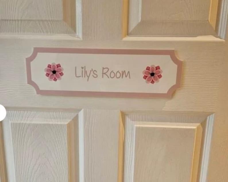 Puede incluir: Un letrero rectangular rosa claro con el texto "Lily's Room" en cursiva. El letrero est&aacute; decorado con dise&ntilde;os florales rosas y rojos. Est&aacute; montado en una puerta blanca.
