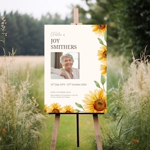 May include: A memorial sign on an easel, featuring a photo of Joy Smithers, with text including dates and event details. The sign is decorated with watercolor sunflowers and green leaves, set against a cream background.