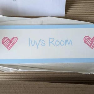 Puede incluir: Un letrero rectangular con fondo blanco y borde azul claro. El letrero presenta el texto "Ivy's Room" en azul claro, con un coraz&oacute;n rosa a cada lado. El letrero es probablemente para la habitaci&oacute;n de un ni&ntilde;o.