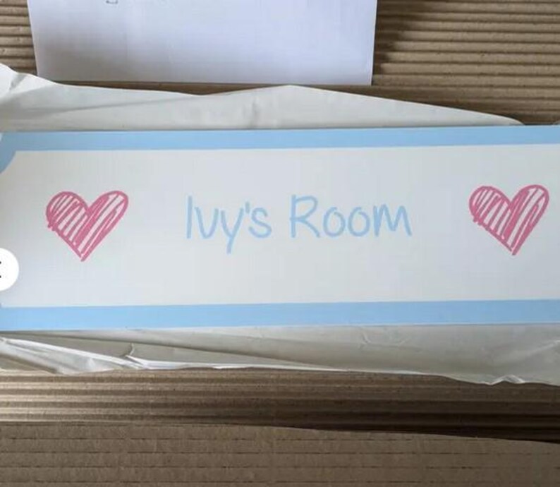 Puede incluir: Un letrero rectangular con fondo blanco y borde azul claro. El letrero presenta el texto "Ivy's Room" en azul claro, con un coraz&oacute;n rosa a cada lado. El letrero es probablemente para la habitaci&oacute;n de un ni&ntilde;o.