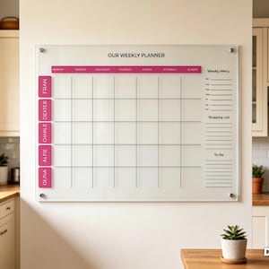 May include: A clear acrylic weekly planner board mounted on a wall with silver hardware. It features a grid with the text OUR WEEKLY PLANNER, days of the week, a weekly menu, a shopping list, a to-do section, and pink labels for names.