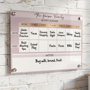 May include: A rectangular acrylic wall-mounted activity planner with a dusty rose background and gold accents. It features a weekly grid labeled The Harper Family, with rows for Jamie and Heidi, columns for days of the week, and a notes section.