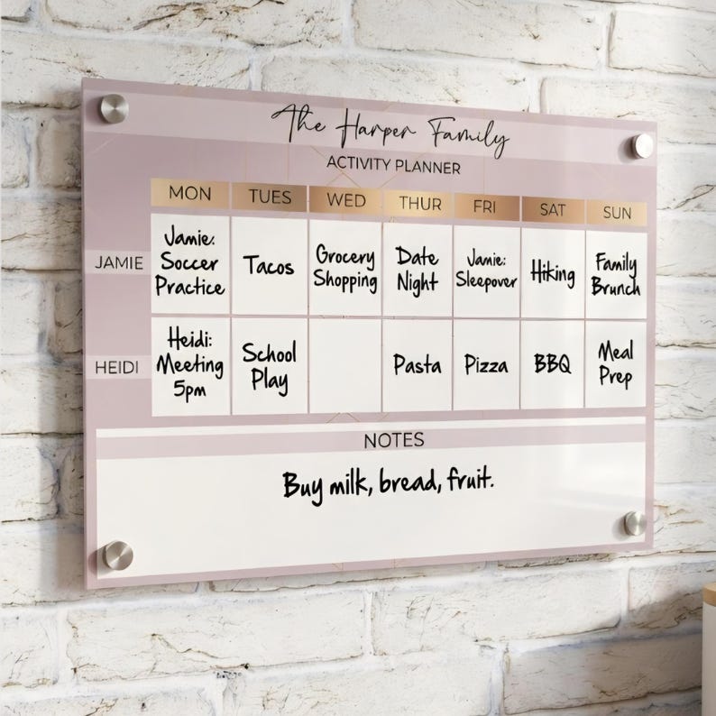 May include: A rectangular acrylic wall-mounted activity planner with a dusty rose background and gold accents. It features a weekly grid labeled The Harper Family, with rows for Jamie and Heidi, columns for days of the week, and a notes section.