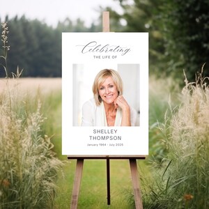 May include: A white sign on a wooden easel with the text "Celebrating the Life of" above a photo of a smiling woman. Below the photo is the name "Shelley Thompson" and the dates "January 1964 - July 2025". The sign is set in a field.