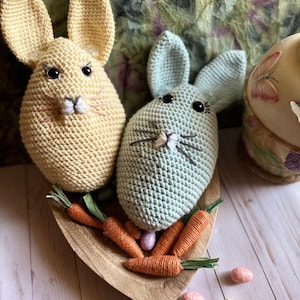 May include: Two crocheted bunny rabbits, one yellow and one light blue, are sitting in a wooden bowl with orange carrots.  There are several colorful Easter eggs scattered on a light wood surface.