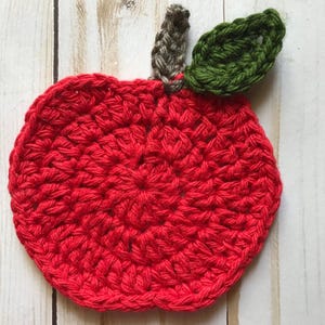 May include: A handmade, red crochet apple coaster with a brown stem and green leaf. The coaster is round and features a textured surface, perfect for protecting tables from hot or cold drinks. The apple design adds a touch of whimsy to any home.