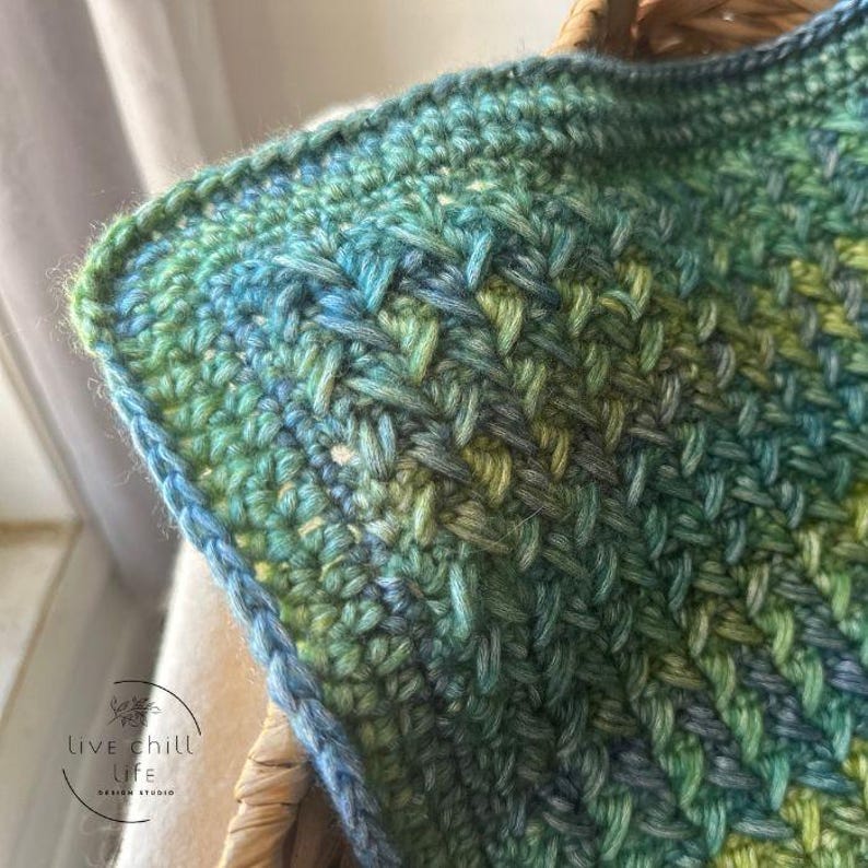 K&ouml;nnte beinhalten: Nahaufnahme einer geh&auml;kelten Decke mit einem Farbverlauf in Gr&uuml;n, Blau und Wei&szlig;. Die Decke hat ein strukturiertes Stichmuster und einen Rand.