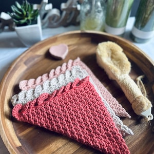 May include: Three crocheted dishcloths in a wooden bowl. The dishcloths are in shades of pink and beige. The top dishcloth is a bright red colour.