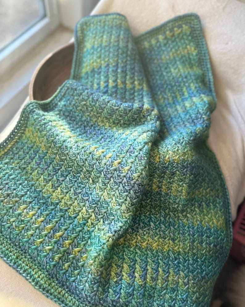 A crocheted blanket in shades of green and blue with a chevron pattern.