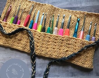 May include: A crocheted roll-up case with a black and gold braided strap, filled with a variety of colorful crochet hooks. The case has a light brown color and is made with a textured stitch. The text "Live Chill Life" is on a round, light blue label with a small leaf design.