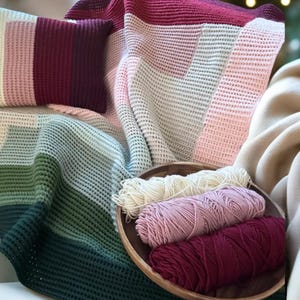 May include: A cozy scene featuring a color-blocked knitted blanket and pillow in shades of pink, white, green, and burgundy. A wooden bowl holds skeins of yarn in complementary colors, suggesting a handmade craft project.