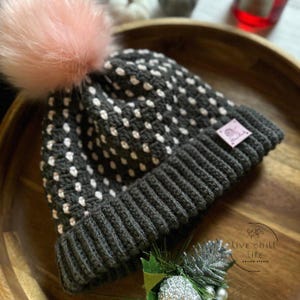 Puede incluir: Un gorro de punto gris oscuro con un estampado de lunares blancos y un pompón rosa. El gorro tiene un borde doblado y una pequeña etiqueta rosa. El texto "live chill life design studio" es visible en la superficie de madera.