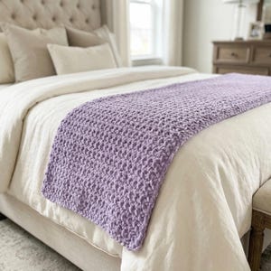 May include: A lavender-coloured, chunky knit throw blanket draped over a bed. The blanket has a textured, woven appearance and is placed on top of a white duvet. The bed has a tufted headboard and several pillows.