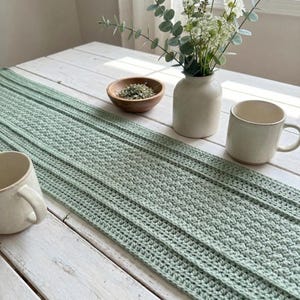 May include: A light green crochet table runner with a textured pattern, displayed on a white wooden table. The runner is paired with a small wooden bowl, a white ceramic vase with flowers, and two white mugs. The scene is bathed in natural light.