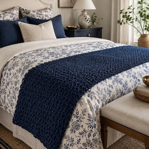May include: A bed featuring a cream headboard and floral bedding. A navy blue knitted throw is draped across the bed. Decorative pillows in navy and cream tones are arranged. A lamp and artwork are also visible.