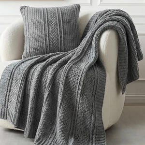 May include: A grey knitted throw and matching cushion are draped over a white armchair. The blanket features a ribbed and patterned design. The cushion has a vertical ribbed pattern. The image text reads: "maker added a variant of border, styled with coordinating pillow pattern also in this shop."