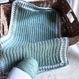 May include: A hand-crocheted blanket in a soft mint green, featuring a ribbed design and a white border. It's displayed in a wicker basket, with a ball of matching yarn nearby. The blanket is likely a small throw or for a baby, showcasing a handmade craft.