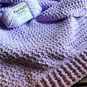 Op de afbeelding: Een lichtpaarse grofgebreide deken gemaakt van zacht chenille-achtig garen. Een enkele bol Premier Parfait Chunky 100% polyestergaren, met een gewicht van 100 gram, rust op het getextureerde, geluste oppervlak van de gevouwen lavendelkleurige stof.