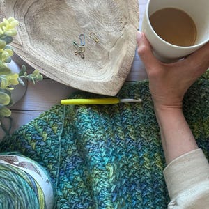 May include: A close-up shot of a crochet project in progress, featuring a vibrant blue and green yarn. A yellow crochet hook rests on the fabric. A wooden heart-shaped bowl holds stitch markers. A cup of coffee is held in the hand.