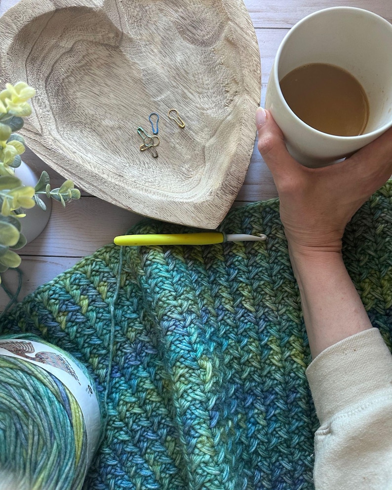 A green, blue, and yellow yarn ball with a label. A crochet hook and a partially completed crochet blanket in green, blue, and yellow colors.