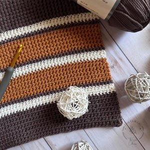 Puede incluir: Una manta de crochet a rayas marrones, blancas y naranjas con una bola de hilo blanca en la parte superior. La manta está hecha con un punto texturizado y tiene un borde de flecos.