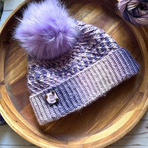 May include: A purple and white crocheted beanie with a fluffy purple pom pom. The beanie has a small, square, wooden tag with a heart and the word "Love" on it.