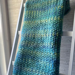 A crocheted blanket in shades of blue and green. The blanket is draped over a white ladder and has a textured, woven pattern.