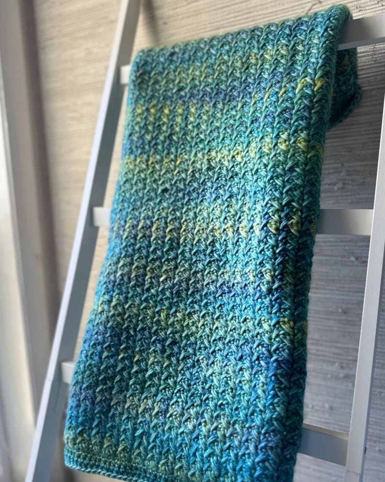 A crocheted blanket in shades of blue and green. The blanket is draped over a white ladder and has a textured, woven pattern.