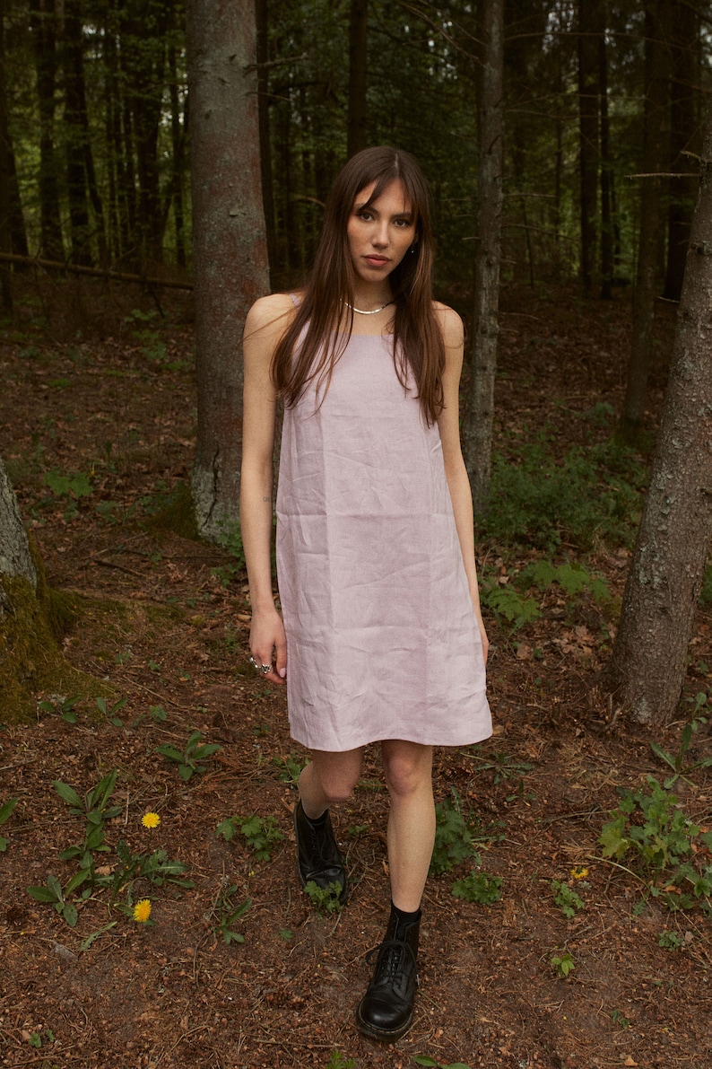 May include: A woman wearing a light pink sleeveless dress and black lace-up boots stands in a forest.