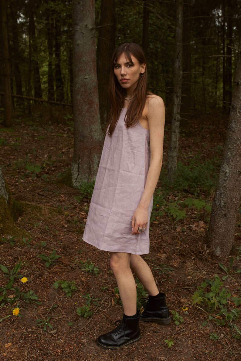 May include: A woman wearing a light purple linen dress and black combat boots stands in a forest.