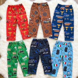 May include: Six pairs of pants featuring various character prints from the Cars movie franchise. The collection includes red, brown, dark blue, green, black, and light blue trousers, each decorated with distinct patterns of race cars, tires, trophies, and text.