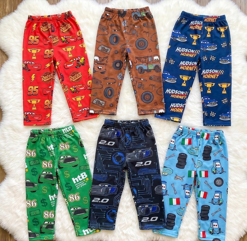 May include: Six pairs of pants featuring various character prints from the Cars movie franchise. The collection includes red, brown, dark blue, green, black, and light blue trousers, each decorated with distinct patterns of race cars, tires, trophies, and text.