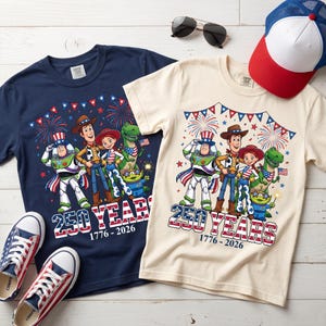 May include: A navy blue and a cream t-shirt featuring a graphic of Toy Story characters wearing patriotic attire with fireworks and bunting. The text reads 250 YEARS 1776-2026. The shirts are displayed with a red, white, and blue baseball cap and sneakers.