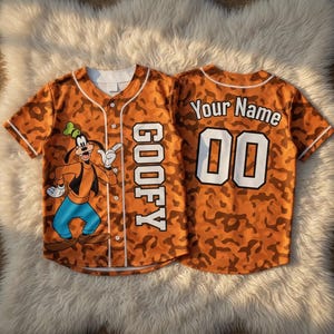 May include: A short-sleeved baseball jersey featuring an orange camouflage pattern. The front displays a graphic of Goofy and the text GOOFY vertically. The back shows the text Your Name above the number 00 in white with black outlines.