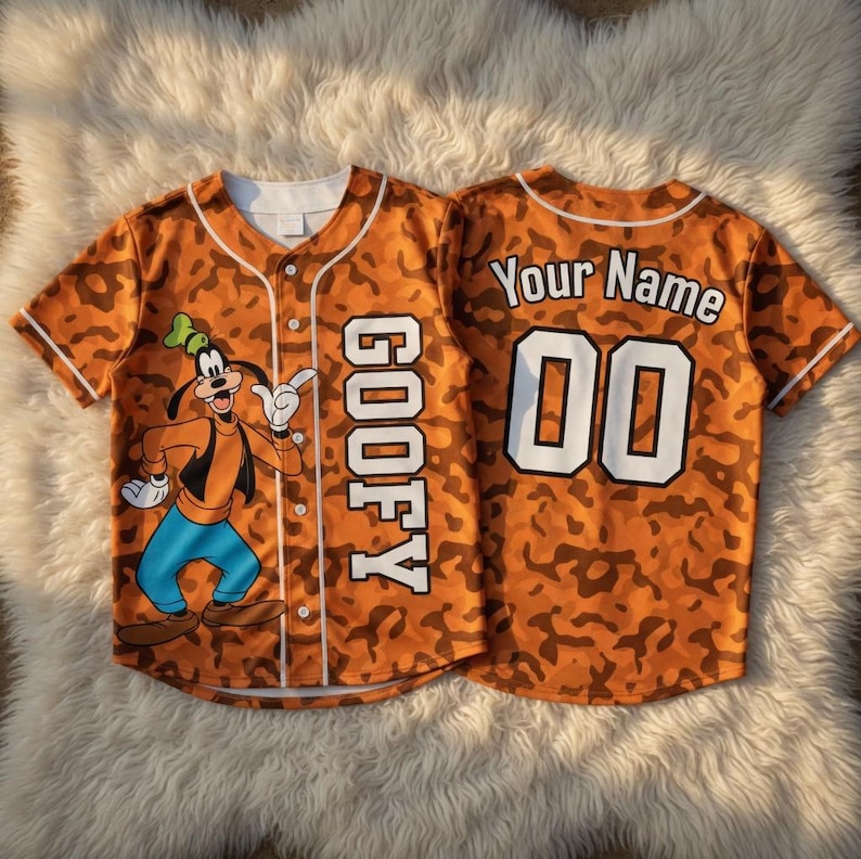 May include: A short-sleeved baseball jersey featuring an orange camouflage pattern. The front displays a graphic of Goofy and the text GOOFY vertically. The back shows the text Your Name above the number 00 in white with black outlines.