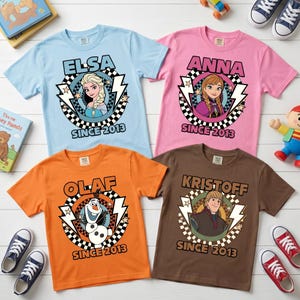 May include: Four short-sleeve t-shirts in light blue, pink, orange, and brown. Each features a circular graphic with a character from Frozen, a checkered border, lightning bolts, and the text "ELSA," "ANNA," "OLAF," or "KRISTOFF" above "SINCE 2013."
