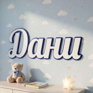 Op de afbeelding: Een wandbord met de naam "Дани" in grote, witte, driedimensionale Cyrillische letters met een donkerblauwe omlijning. Het bord is bevestigd aan een lichtblauwe muur met een patroon van witte wolken en kleine sterren.
