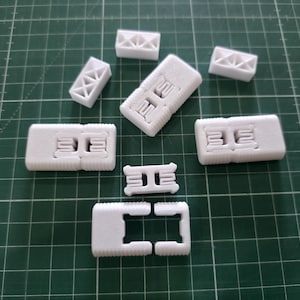 Könnte beinhalten: Eine Sammlung weißer, 3D-gedruckter Kunststoffkomponenten mit rechteckigen Formen, internen Gitterstrukturen, ineinandergreifenden Mechanismen und texturierten Kanten, die verstreut auf einer grünen Schneidematte mit Raster angeordnet sind.