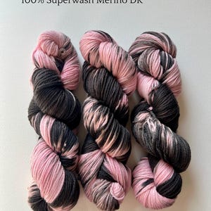 以下が含まれることがあります： 「Strawberry Cow」というカラーウェイの3つの糸玉。ピンクと濃いグレーの混紡糸で、「100% Superwash Merino DK」の文字が見えます。白い背景に糸が並んでいます。