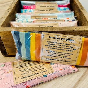 May include: A wooden box filled with colorful lavender eye pillows. The pillows are made of fabric with different patterns and are packaged in clear plastic. The box has a label that says "lavender eye pillow" and includes instructions for use. The label also says "handmade with love & kisses!"