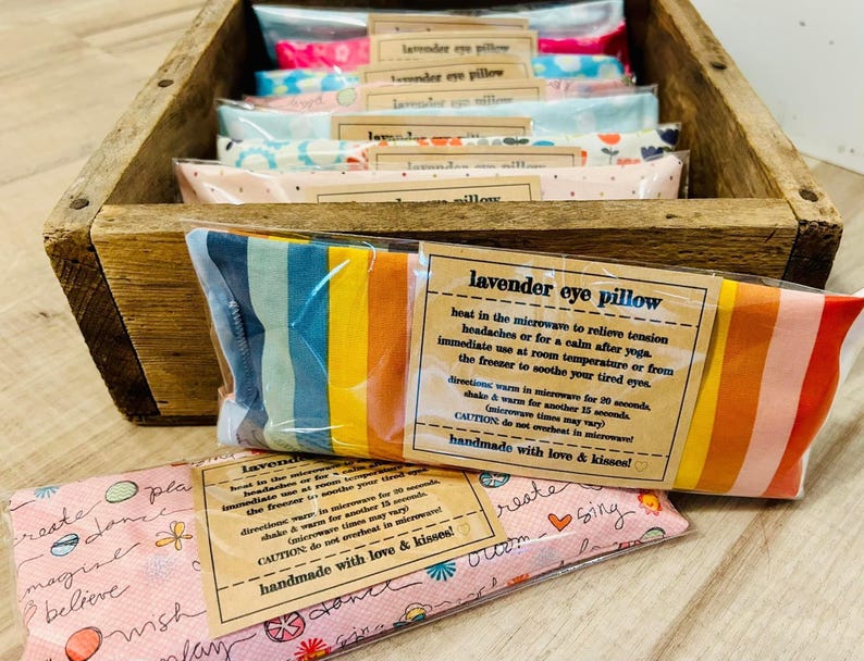 May include: A wooden box filled with colorful lavender eye pillows. The pillows are made of fabric with different patterns and are packaged in clear plastic. The box has a label that says "lavender eye pillow" and includes instructions for use. The label also says "handmade with love & kisses!"