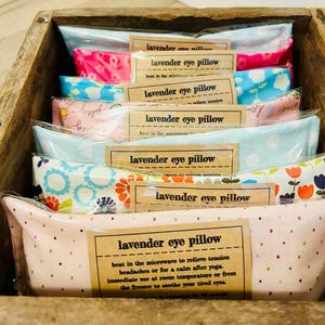 May include: A stack of lavender eye pillows in a wooden crate. The pillows are made of different fabrics with floral patterns and are labeled "lavender eye pillow". The text on the label says "heat in the microwave to relieve tension, headaches or for a calm after yoga. Immediate use at room temperature or from the freezer to soothe your tired eyes."