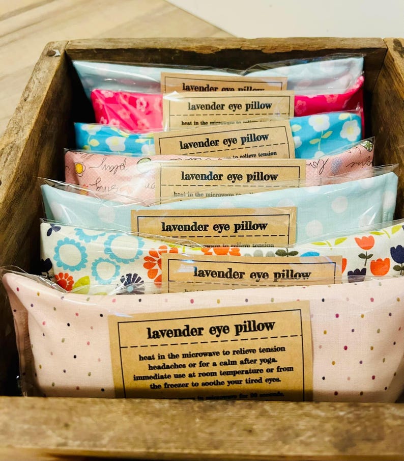 May include: A stack of lavender eye pillows in a wooden crate. The pillows are made of different fabrics with floral patterns and are labeled "lavender eye pillow". The text on the label says "heat in the microwave to relieve tension, headaches or for a calm after yoga. Immediate use at room temperature or from the freezer to soothe your tired eyes."