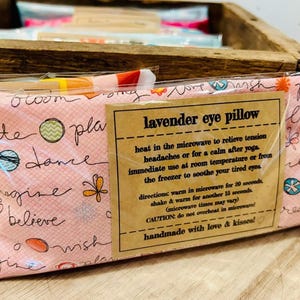 May include: A pink lavender eye pillow with a white and black floral pattern and the text "lavender eye pillow" on a brown label. The label also includes instructions for using the pillow to relieve tension headaches, calm after yoga, or soothe tired eyes.