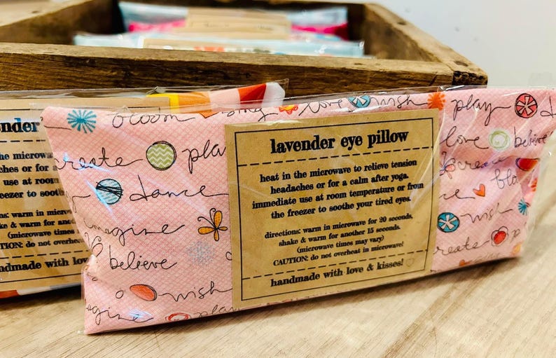 May include: A pink lavender eye pillow with a white and black floral pattern and the text "lavender eye pillow" on a brown label. The label also includes instructions for using the pillow to relieve tension headaches, calm after yoga, or soothe tired eyes.